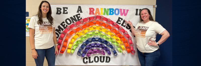 Counselors Rebecca Chadick and Courtney Taylor in front of a rainbow chain link poster